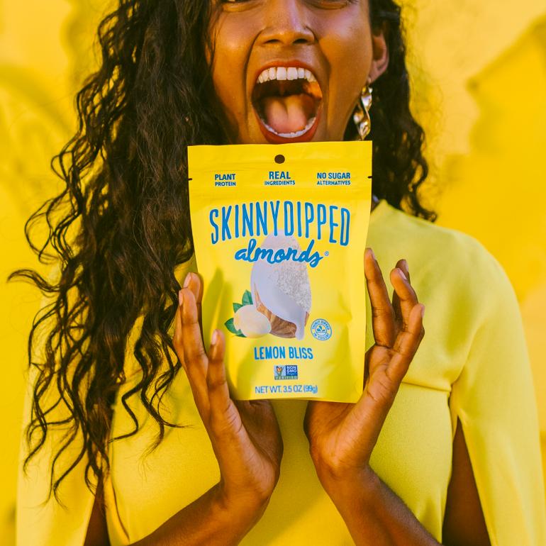 Girl in a yellow dress holding a bag of SkinnyDipped Lemon Bliss Almonds on a yellow background.